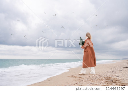 Blond woman Christmas tree sea. Christmas portrait of a happy woman walking along the beach and holding a Christmas tree on her shoulder. She is wearing a brown coat and a white suit. 114463297