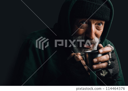 Old homeless man with grey beard holding a mug of hot tea to warm himself. 114464376