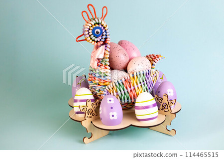 A rainbow chicken woven from a paper vine with Easter eggs inside. It stands on a wooden stand with eggs on a blue background. The concept of Easter 114465515