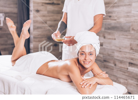 client smiling a nd looking at the camera while her masseur holding a bowl of oil 114465671