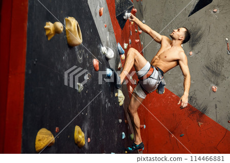 Male climber exercising in gym, climbing up on artificial wall with boulders. Male climber exercising in gym, climbing up on artificial wall with boulders. 114466881