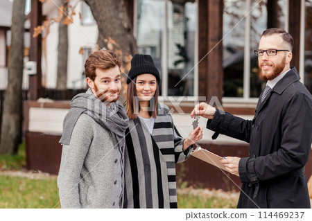 Friendly male real estate agent handing house key to a smiling young couple in office Friendly male real estate agent handing house key to a smiling young couple in office 114469237