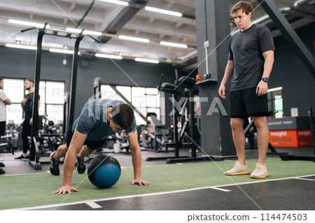 Professional sport coach explaining to beginner sportsman how to training with heavy fitness ball at gym. Motivated young man working out with medicine ball support under supervision of trainer. 114474503