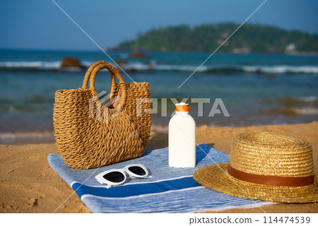 Straw bag, white sunscreen bottle, hat, sunglasses on blue towel at sunny beach. Holiday items for skin protection, sunbathing by the sea. Ocean, rocks, island background on clear day 114474539
