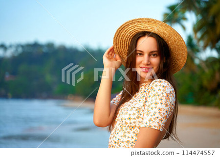 Smiling woman in floral dress holds straw hat on tropical beach at sunset. Happy female enjoys summer evening by sea, palm trees in background. Traveler poses during golden hour on island coast. 114474557