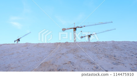 Three construction cranes tower over a mound of sand against a backdrop of blue sky. Perfect for illustrating construction progress and industrial development Three construction cranes tower over a mound of sand against a backdrop of blue sky. Perfect for illustrating construction progress and industrial development 114475910