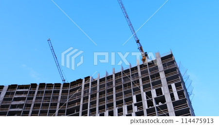 Two construction cranes tower over a multi-story building under concrete construction, windows yet to be installed, against a blue sky backdrop. Ideal for showcasing urban development 114475913