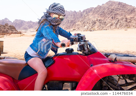 Brave kid in checkered scarf around head is riding red quad bike in sand desert Brave kid in checkered scarf around head is riding red quad bike in sand desert 114476034