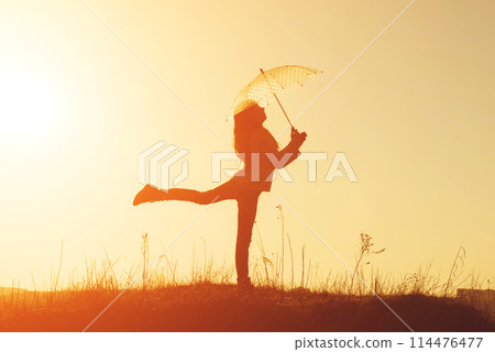 Dreamy little girl standing on one leg with small umbrella at sunset. Silhouette of happy girl with umbrella Dreamy little girl standing on one leg with small umbrella at sunset. Silhouette of happy girl with umbrella 114476477