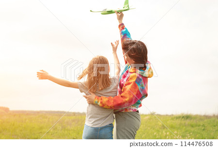 Mother and daughter launching toy plane at the field. Girl and woman spending family time together at the nature. Portrait from back with sunset rays 114476508