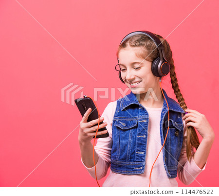 Happy teenage girl listening to music in big headphones while playing with smartphone on pink background Happy teenage girl listening to music in big headphones while playing with smartphone on pink background 114476522