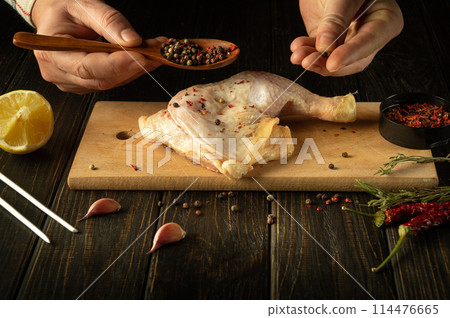 Close-up of a chef hands with a spoon adding pepper to a chicken leg. Broiler leg on cutting board with rosemary and spices before grilling Close-up of a chef hands with a spoon adding pepper to a chicken leg. Broiler leg on cutting board with rosemary and spices before grilling 114476665