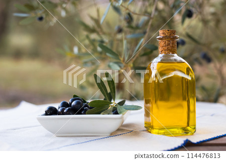 Olive oil in a glass bottle and green olives on the background of olive branches in the garden Olive oil in a glass bottle and green olives on the background of olive branches in the garden 114476813