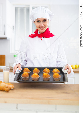 Proffesional woman cook in white uniform holding sheet pan with just baked cupcakes Proffesional woman cook in white uniform holding sheet pan with just baked cupcakes 114476816