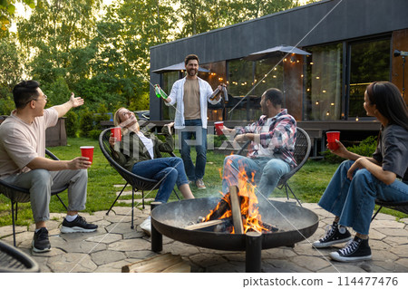 Cheerful multicultural friends sitting near fire on backyard enjoying happy weekend 114477476