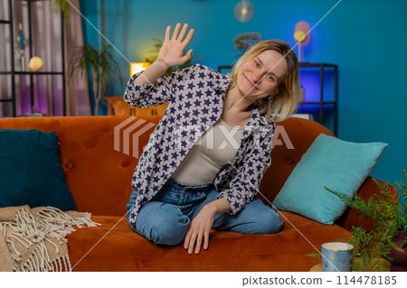 Portrait of young woman smiling at camera and waving gesturing hello hi greeting or goodbye at home 114478185