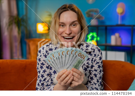 Happy excited girl winning online casino lottery game waving dollar bill money fan sitting on couch 114478194