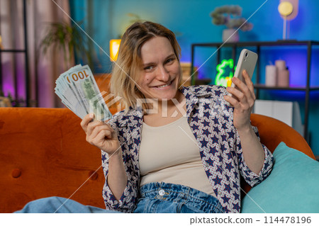 Successful rich smiling woman holding smartphone waving dollar bill money fan sitting on home sofa 114478196