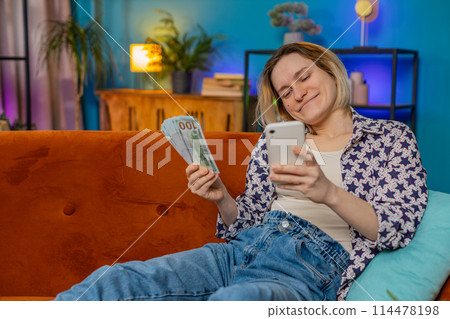 Successful rich smiling woman holding smartphone waving dollar bill money fan sitting on home sofa Successful rich smiling woman holding smartphone waving dollar bill money fan sitting on home sofa 114478198