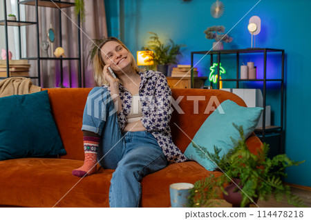 Happy young Caucasian woman making mobile phone conversation with friends sitting on couch at home 114478218