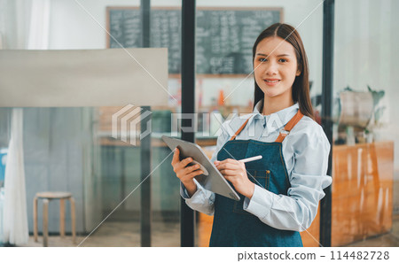 Smiling Asian coffee shop owner standing in front of cashier counter with tablet ready to take menu orders.welcome customers who come in to place orders in the cafe. 114482728