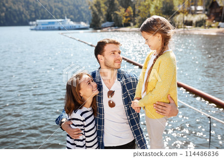 Happy family spending time together outside near lake. Parents playing with daughter hugging and having fun 114486836