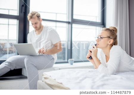 man is working on the laptop at home while his wife resting on the bed man is working on the laptop at home while his wife resting on the bed 114487879