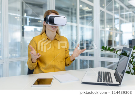 Engaged young female professional using VR headset to participate in a virtual meeting from a bright office environment. Engaged young female professional using VR headset to participate in a virtual meeting from a bright office environment. 114489308