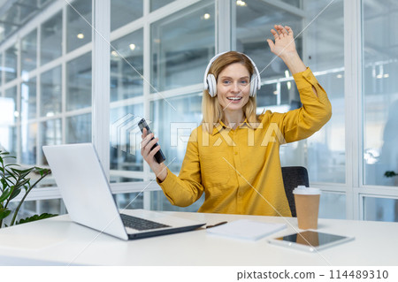 A joyful female professional in a vibrant yellow shirt waves while enjoying music on headphones, showcasing work-life balance in a modern office setting. 114489310