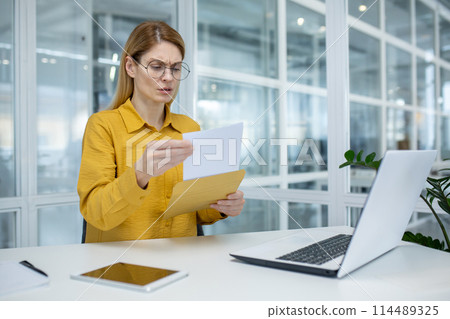 A focused businesswoman in a modern office reading documents from a yellow envelope, displaying a confused and bothered expression. A focused businesswoman in a modern office reading documents from a yellow envelope, displaying a confused and bothered expression. 114489325
