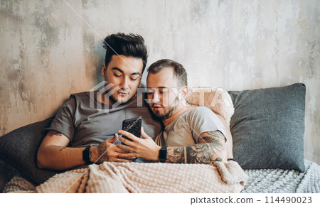 Happy gay couple lying together in bed and using smartphone, looking at photos Happy gay couple lying together in bed and using smartphone, looking at photos 114490023