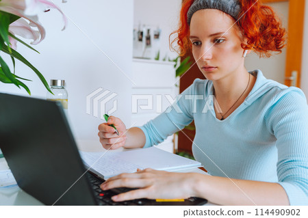 portrait red-haired teenage girl sitting at table in room using computer 114490902