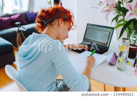 portrait red-haired teenage girl sitting at table in room using computer portrait red-haired teenage girl sitting at table in room using computer 114490922