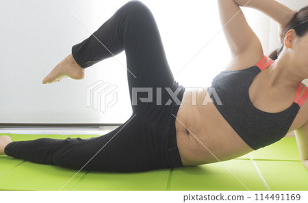 A woman doing side crunches on a yoga mat with stretch marks on her postpartum belly 114491169