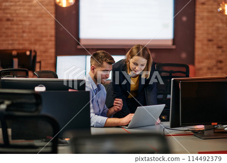 Business colleagues, a man and a woman, engage in discussing business strategies while attentively gazing at a computer monitor, epitomizing collaboration and innovation 114492379