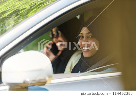 Two Muslim women wearing hijab converse on a smartphone while traveling together in a car through the  114492393