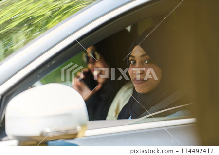 Two Muslim women wearing hijab converse on a smartphone while traveling together in a car through the  114492412