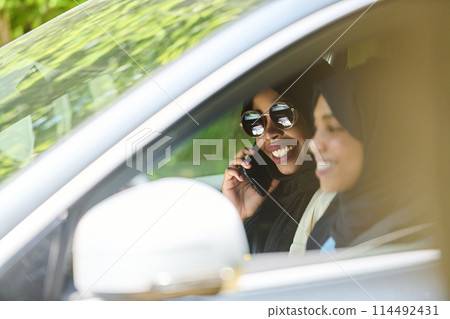 Two Muslim women wearing hijab converse on a smartphone while traveling together in a car through the  114492431