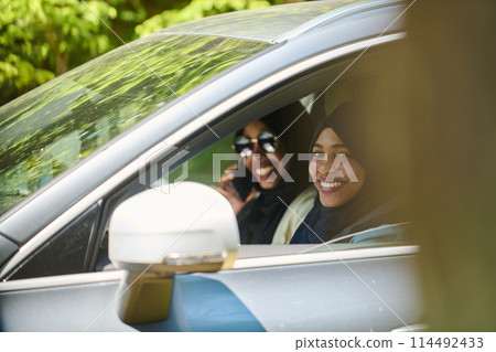 Two Muslim women wearing hijab converse on a smartphone while traveling together in a car through the  114492433