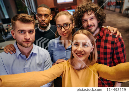 A diverse group of business professionals take a break from their tasks in a modern startup office to capture a creative selfie, showcasing teamwork and a vibrant workplace culture 114492559