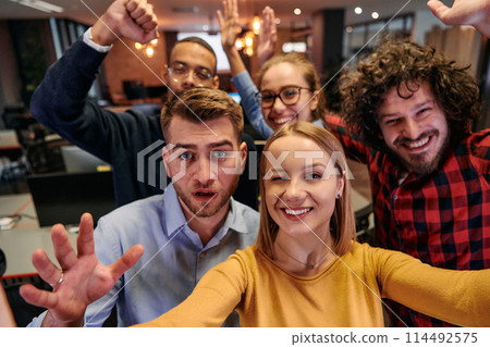 A diverse group of business professionals take a break from their tasks in a modern startup office to capture a creative selfie, showcasing teamwork and a vibrant workplace culture 114492575