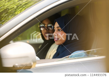 Two Muslim women wearing hijab converse on a smartphone while traveling together in a car through the  114493376