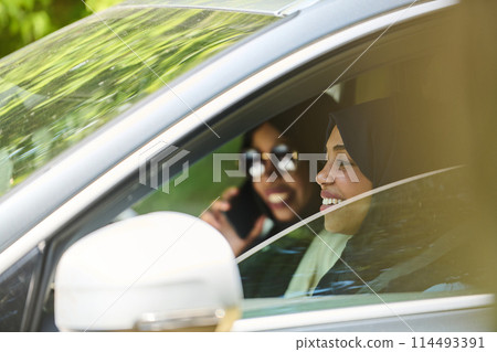 Two Muslim women wearing hijab converse on a smartphone while traveling together in a car through the  114493391