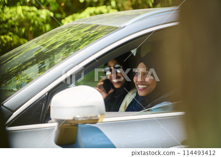Two Muslim women wearing hijab converse on a smartphone while traveling together in a car through the Two Muslim women wearing hijab converse on a smartphone while traveling together in a car through the 114493412