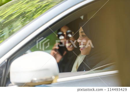 Two Muslim women wearing hijab converse on a smartphone while traveling together in a car through the  114493429