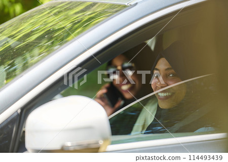 Two Muslim women wearing hijab converse on a smartphone while traveling together in a car through the  114493439