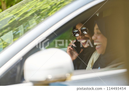 Two Muslim women wearing hijab converse on a smartphone while traveling together in a car through the  114493454