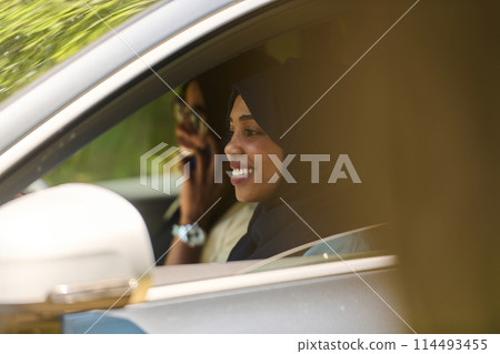 Two Muslim women wearing hijab converse on a smartphone while traveling together in a car through the  114493455