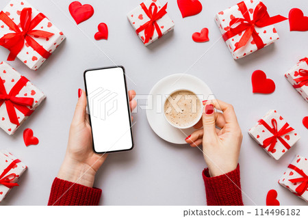 Female hands holding smart phone with coffee for Valentine day, gift box and envelope, hearts 114496182