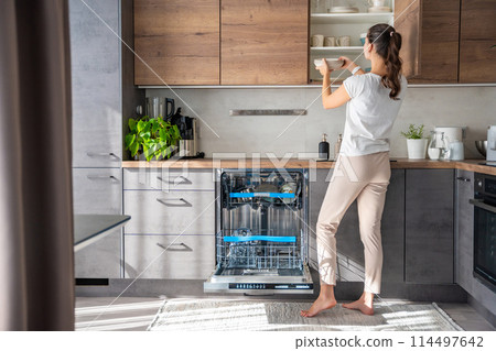 Young woman removes clean ceramic dishes from the dishwasher. Household and useful technology concept. Young woman removes clean ceramic dishes from the dishwasher. Household and useful technology concept. 114497642